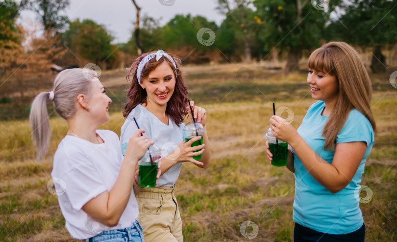 Скачать Три молодые женщины в модной одежде, за городом, на природе, летом и пьют освежающие безалкогольные коктейли из одноразовых пластиковых стаканчиков. Женская дружба. Каникулы, приятный досуг. фотосток Ozero