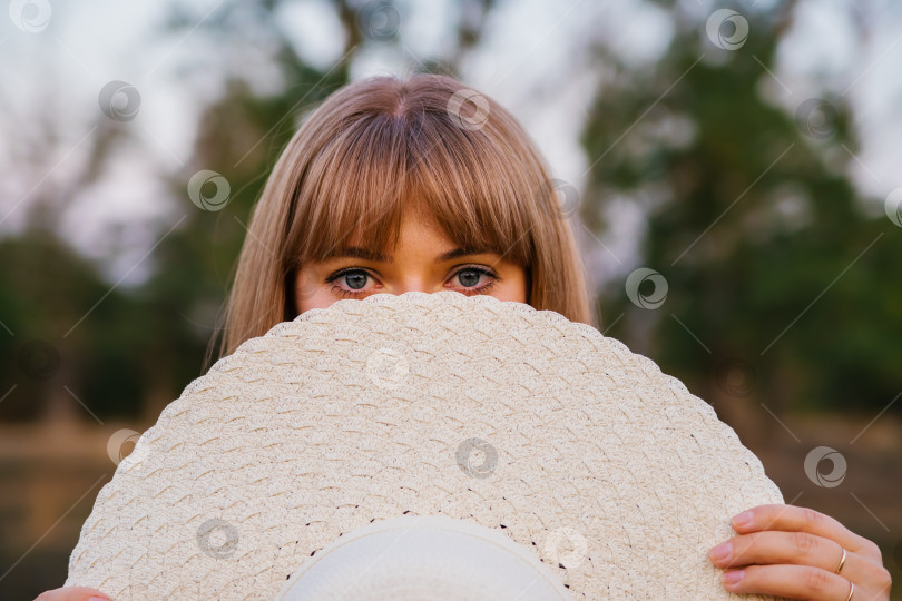 Скачать Молодая женщина закрыла лицо шляпой. Красивые глаза девушки видны из-под шляпы. Соломенная летняя шляпа, модный аксессуар. Концепция летних каникул. фотосток Ozero