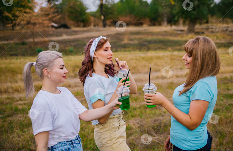 Скачать Три молодые женщины в модной одежде, за городом, на природе, летом и пьют освежающие безалкогольные коктейли из одноразовых пластиковых стаканчиков. Женская дружба. Каникулы, приятный досуг. фотосток Ozero