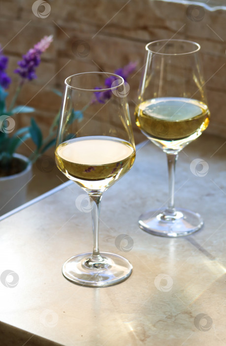 Скачать Два бокала белого вина на кухонном столе перед окном с фиолетовым цветком на обратной стороне в солнечный день. Крупный план сверкающих бокалов с белым вином на столе при естественном освещении фотосток Ozero