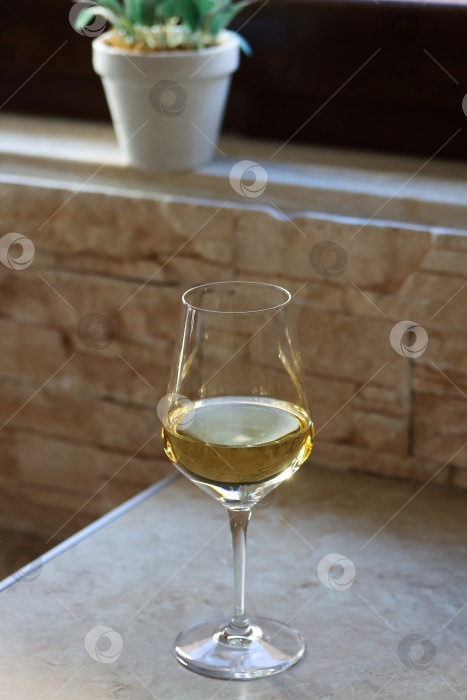 Скачать Бокал белого вина на кухонном столе перед окном с цветком на обратной стороне в солнечный день. Крупный план блестящего бокала белого вина на столе с местом для копирования текста фотосток Ozero