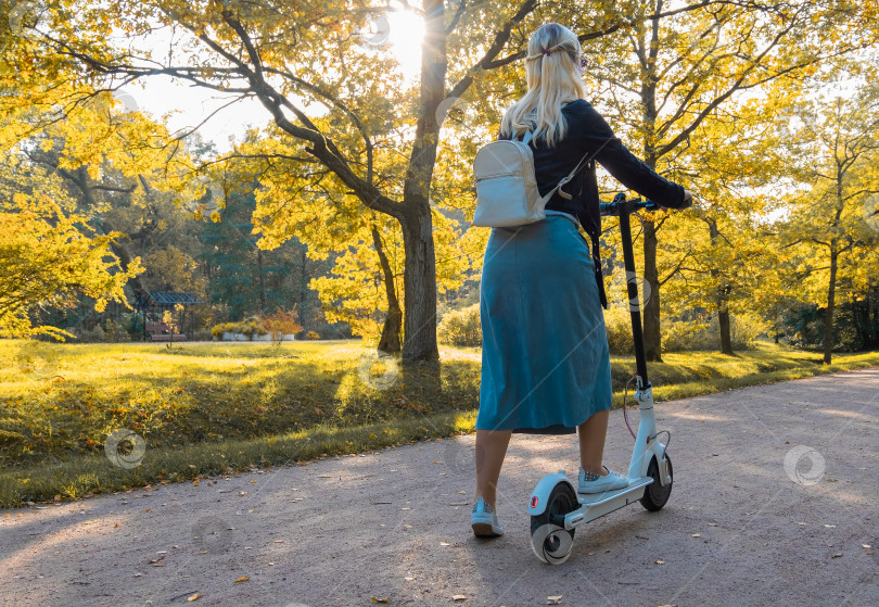 Скачать Молодая блондинка с рюкзаком собирается прокатиться на электрическом скутере из парка ранней осенью. фотосток Ozero