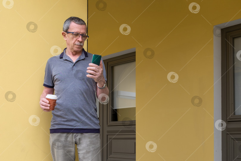 Скачать Взрослый пенсионер стоит и держит смартфон и стакан кофе с собой на выход фотосток Ozero