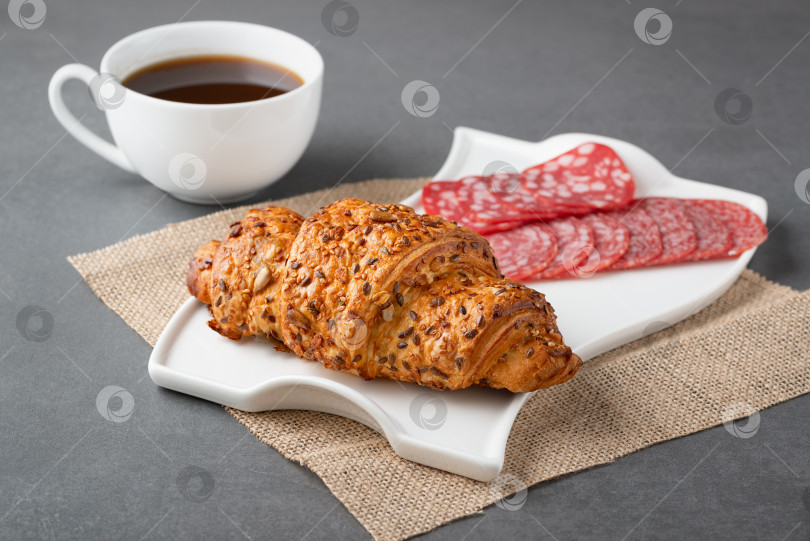 Скачать Свежий бутерброд с круассаном, колбасой и кофе на сером столе фотосток Ozero