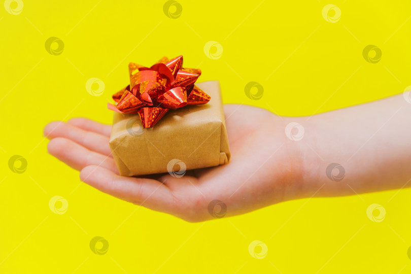 Скачать Детская рука держит маленькую подарочную коробку с красным бантом на желтом фоне. Подарок на день рождения. фотосток Ozero