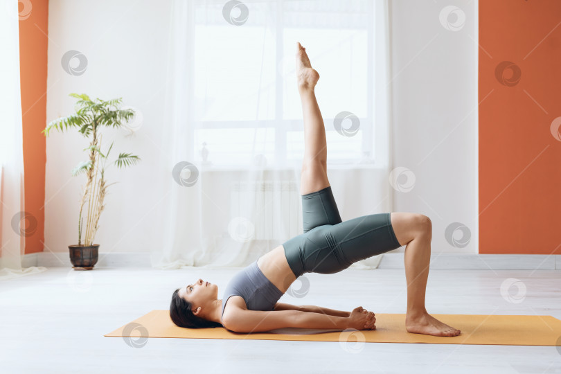 Скачать Молодая женщина в леггинсах и короткой футболке, практикующая йогу, выполняет двипада питхасану с вытянутой вверх ногой, поза полумоста фотосток Ozero