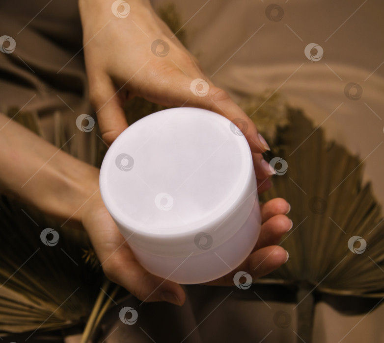 Скачать Женские руки держат белую баночку с кремом. Пластиковый контейнер с косметикой на фоне бежевой ткани. фотосток Ozero