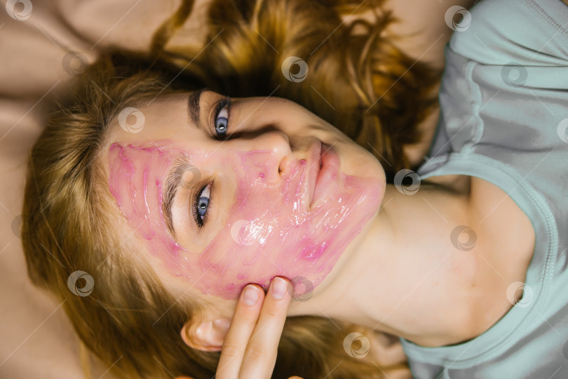 Скачать Молодая женщина наносит на лицо розовую косметическую маску или гель. Уход за кожей лица. фотосток Ozero