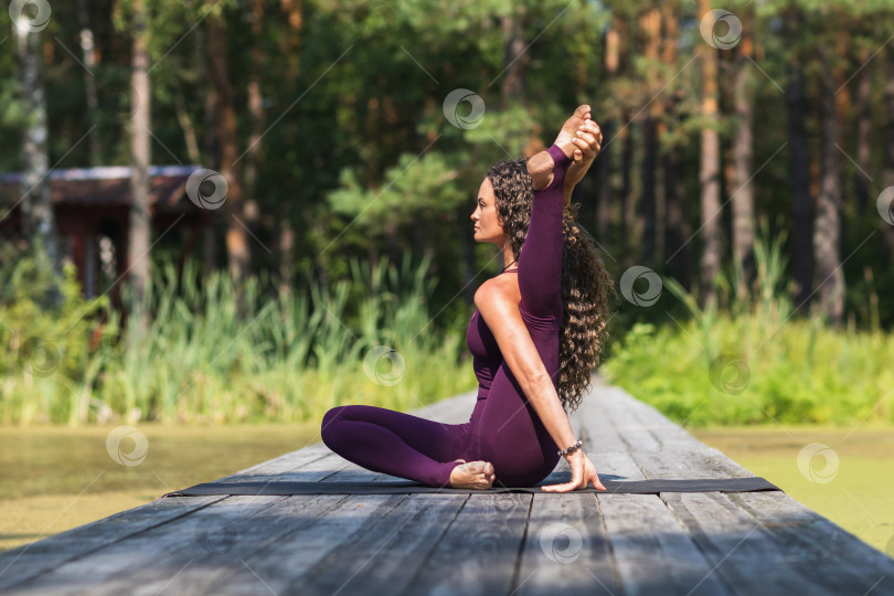 Скачать Женщина, ведущая здоровый образ жизни и практикующая йогу, выполняет вариацию упражнения янтрасана, позу компаса, тренируется в спортивной одежде в парке, сидя на деревянном мосту фотосток Ozero