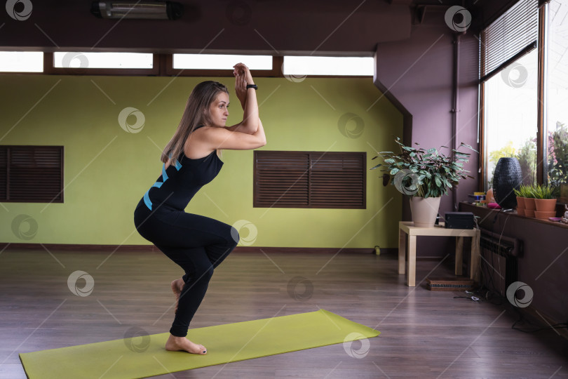 Скачать Женщина, ведущая здоровый образ жизни и практикующая йогу, выполняет упражнение гарудасана, позу орла, тренируется в комнате, стоя на коврике фотосток Ozero