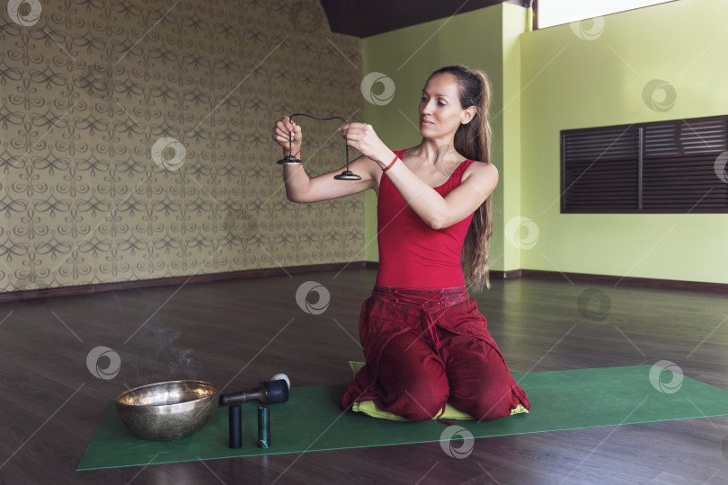 Скачать Женщина, практикующая йогу, занимающаяся звуковой медитацией с колокольчиками, сидит на коврике в комнате фотосток Ozero