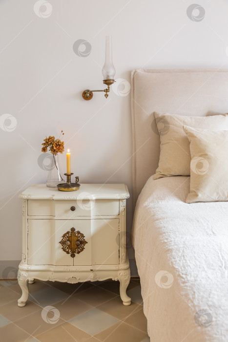 Скачать Классическая спальня с деревянной прикроватной тумбочкой с горящей свечой и цветами возле удобной кровати. Интерьер уютного дома в стиле ретро. фотосток Ozero