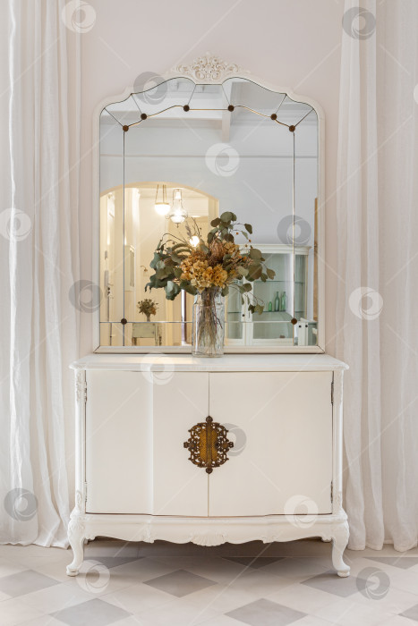 Скачать Старинная отреставрированная мебель в классическом светлом интерьере с белыми стенами и шторами. Винтажное зеркало и букет цветов на деревянном шкафу. фотосток Ozero