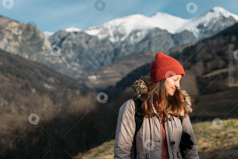 Скачать Портрет улыбающегося путешественника на фоне заснеженных гор. Счастливая женщина в красной шапочке, согревающаяся и наслаждающаяся солнечным светом во время пешего похода в горы. фотосток Ozero
