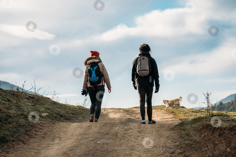 Скачать Молодая пара с рюкзаками и собакой гуляет в горах. Мужчина и женщина, тепло одетые, идут по пешеходной тропе. фотосток Ozero