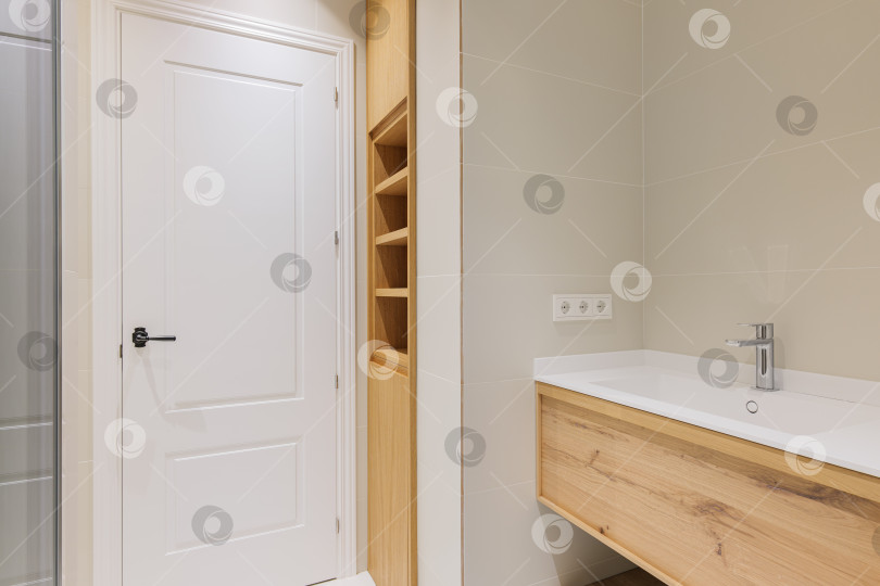Скачать Современная ванная комната с деревянным основанием для белой раковины, отделанная светлой плиткой. Встроенная деревянная мебель с полками. фотосток Ozero