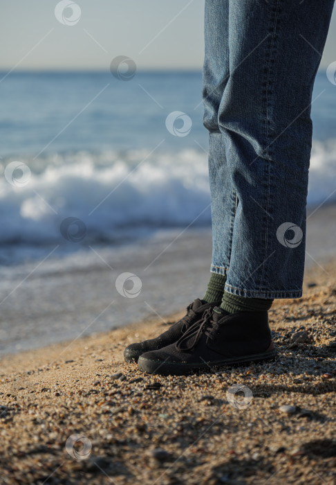Скачать Ноги в синих джинсах и черных туфлях стоят на пляже на фоне морских волн фотосток Ozero