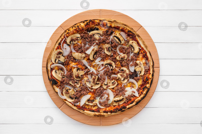 Скачать Цельнокруглая пицца на деревянной тарелке, посыпанная грибами, луком и рубленым мясом. Вид сверху на фоне белой доски фотосток Ozero