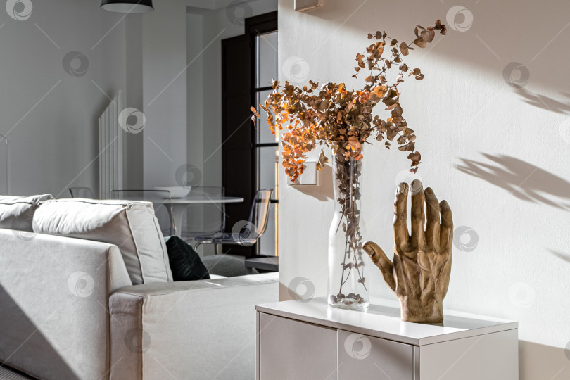 Скачать Декоративная ручная скульптура и ваза с цветами на маленьком белом шкафчике в солнечном интерьере современной квартиры фотосток Ozero