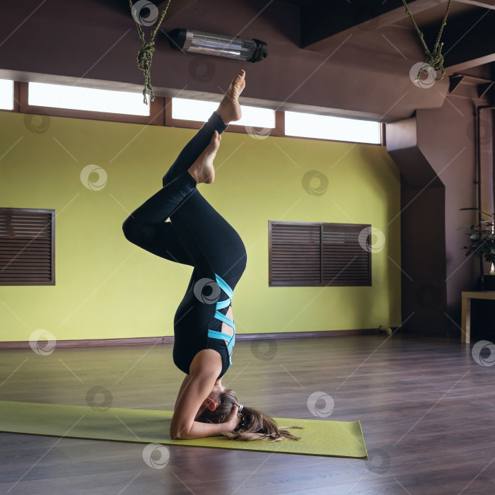 Скачать Женщина, практикующая йогу, выполняет вариацию упражнения ширшасана, перевернутую асану, тренируется на коврике в студии фотосток Ozero