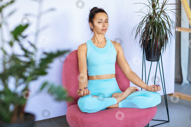 Скачать Женщина в синей спортивной одежде занимается йогой, сидит в позе лотоса на стуле и медитирует с закрытыми глазами фотосток Ozero