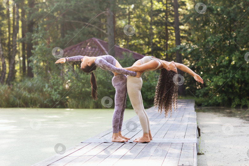 Скачать Две практикующие йогу женщины, держась друг за друга, выполняют Хаста Уттанасану, позу прогиба спины, стоя на деревянном пешеходном мостике в парке фотосток Ozero