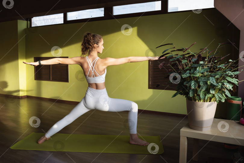 Скачать Молодая привлекательная женщина, практикующая йогу, выполняющая упражнение Вирабхадрасана, поза воина, тренирующаяся в белой спортивной одежде в комнате на коврике, стоя спиной к камере фотосток Ozero