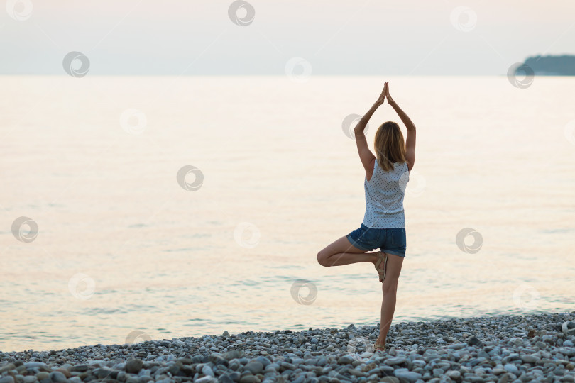 Скачать Женщина, практикующая йогу на берегу моря, выполняет упражнение врикшасана, позу дерева фотосток Ozero