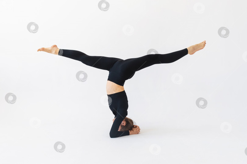 Скачать Женщина в черной спортивной одежде, практикующая йогу, выполняет упражнения ширшасана с хануманасаной, стойка на голове фотосток Ozero