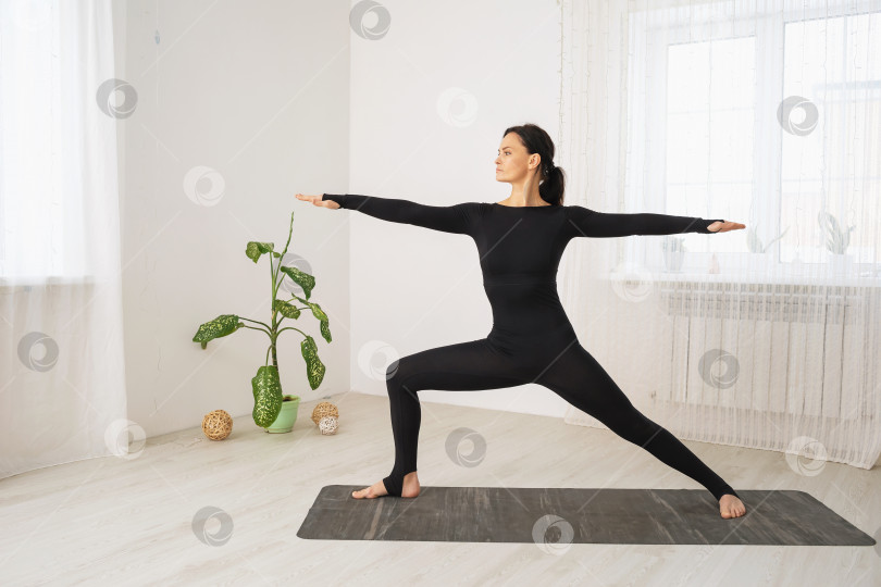 Скачать Женщина в черной спортивной одежде, практикующая йогу, выполняющая упражнение Вирабхадрасана, первую позу воина, стоит на коврике в комнате у окна фотосток Ozero