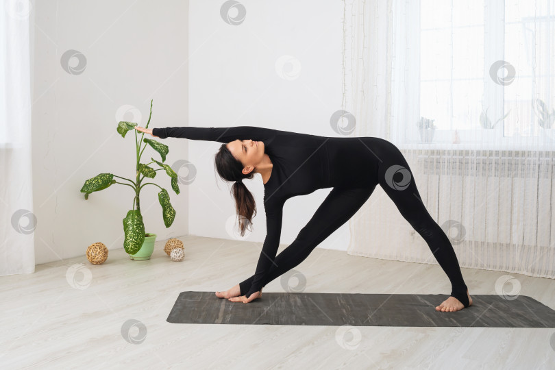 Скачать Женщина в черном спортивном комбинезоне, занимающаяся йогой, выполняет вариацию упражнения Уттхита триконасана, позу вытянутого треугольника, тренируясь стоя на коврике в комнате фотосток Ozero