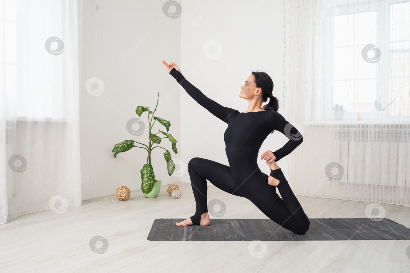 Скачать Женщина в черной спортивной одежде, практикующая йогу, выполняет упражнение анджанейасана с вирасаной, поза низкого выпада, сидя на коврике в комнате у окна фотосток Ozero