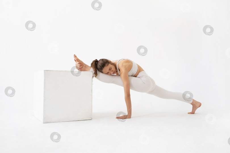 Скачать Молодая женщина, ведущая здоровый образ жизни и практикующая йогу, выполняет упражнение Хануманасана, продольный шпагат, тренируется в белой спортивной одежде в студии на светлом фоне фотосток Ozero
