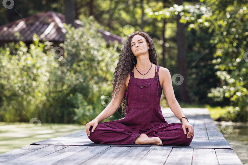 Скачать Женщина, практикующая йогу, медитирующая с закрытыми глазами, сидя в позе лотоса на деревянном мосту, на берегу пруда теплым солнечным утром фотосток Ozero