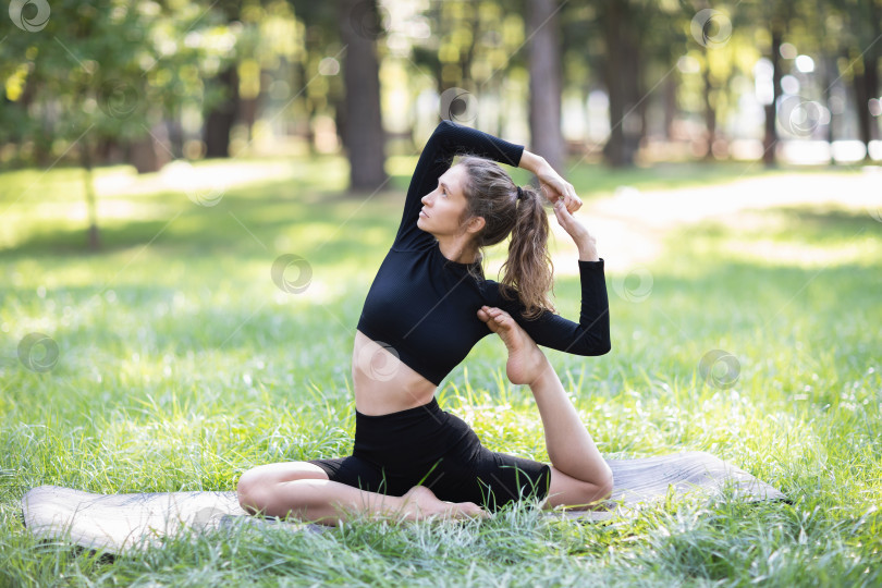 Скачать Молодая женщина в черной спортивной одежде, практикующая йогу, выполняет вариацию упражнения Эка Пада Раджакапотасана, поза голубя, тренируется летним утром в парке, сидя на коврике фотосток Ozero