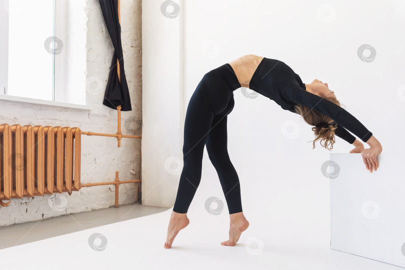 Скачать Молодая женщина, практикующая йогу, выполняет упражнение Урдхва Дханурасана, позу моста из положения стоя, опираясь руками на куб, стоит в светлой студии у окна фотосток Ozero