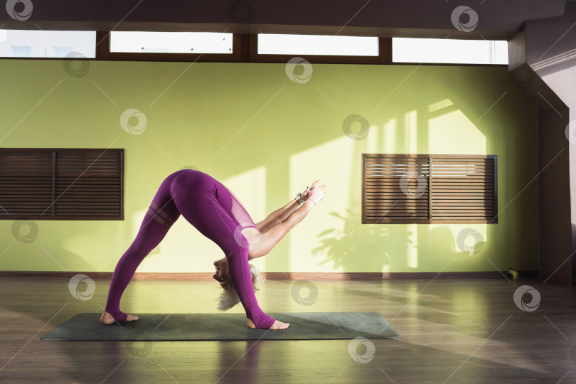 Скачать Женщина, ведущая здоровый образ жизни и практикующая йогу, выполняет упражнение прасарита падоттанасана, наклоняется вперед с шагом, тренируется в бордовой спортивной одежде, стоит на коврике в студии фотосток Ozero