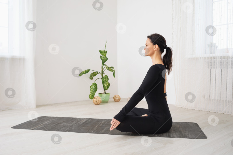 Скачать Женщина в черной спортивной одежде, практикующая йогу, сидит на коврике в позе лотоса, занимается медитацией в комнате, вид сбоку фотосток Ozero