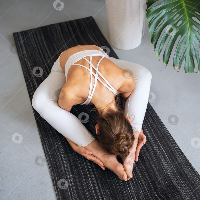 Скачать Молодая спортивная женщина, практикующая йогу, выполняет вариацию упражнения курмасана, позу черепахи, тренируется в одиночестве, сидя на коврике в белой спортивной одежде в комнате фотосток Ozero