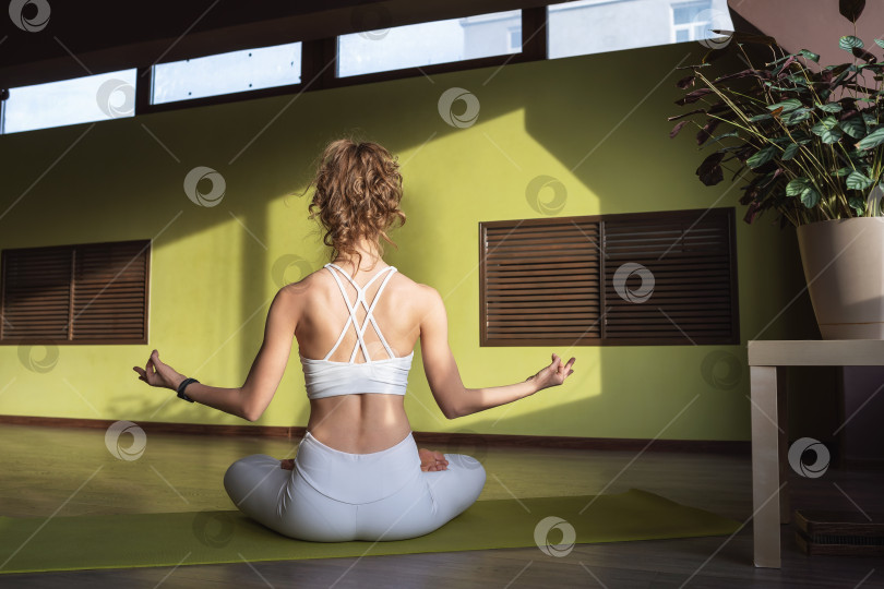 Скачать Молодая женщина, практикующая йогу, медитирующая, сидя в позе лотоса с джняна мудрой, тренирующаяся в белой спортивной одежде в комнате, сидя спиной к камере фотосток Ozero