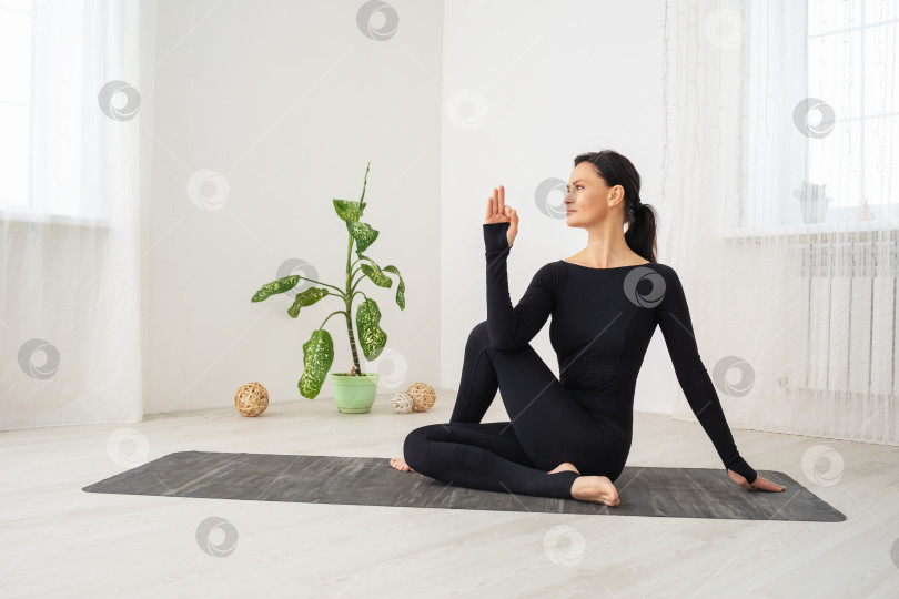 Скачать Женщина в черной спортивной одежде, практикующая йогу, выполняет упражнение Ардха Матсиендрасана, поза короля рыб, тренируется, сидя на коврике в комнате у окна фотосток Ozero