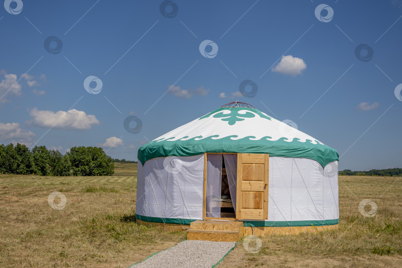 Скачать Белая башкирская юрта (дом кочевых народов) стоит в чистом поле на фоне голубого летнего облачного неба. Дверь юрты открыта и к ней ведет гравийная дорожка. фотосток Ozero