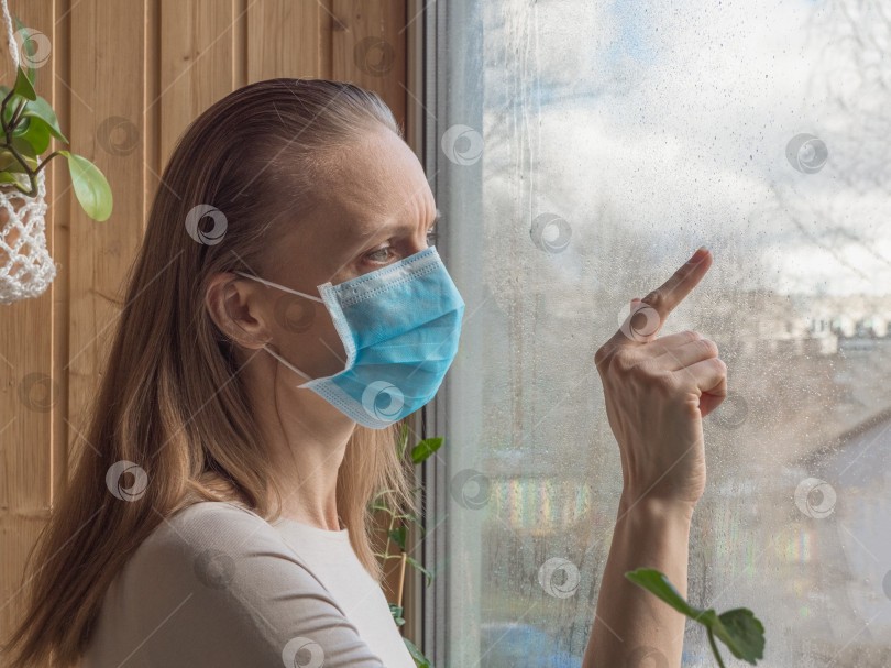 Скачать Карантин, пандемия коронавируса. Женщина в медицинской одноразовой маске печально стоит у окна фотосток Ozero