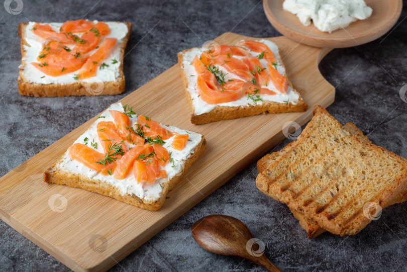 Скачать Домашний хлеб на деревянной разделочной доске с творожным сыром, лососем и зеленью. Украшен зеленью фотосток Ozero
