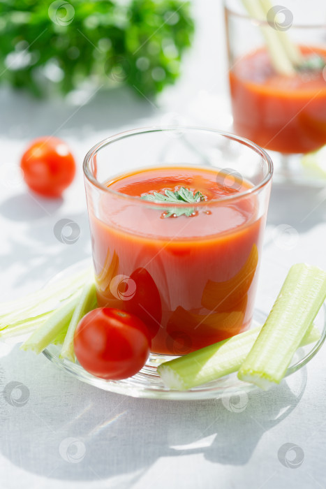 Скачать Свежий томатный сок. Томатный сок наливают в прозрачный стакан со стеблями петрушки и сельдерея на белом фоне, освещенном солнечным светом фотосток Ozero