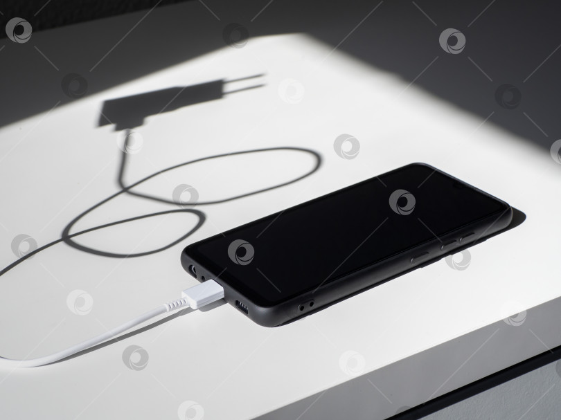 Скачать Черный смартфон с зарядным проводом на белом столе с тенями. фотосток Ozero