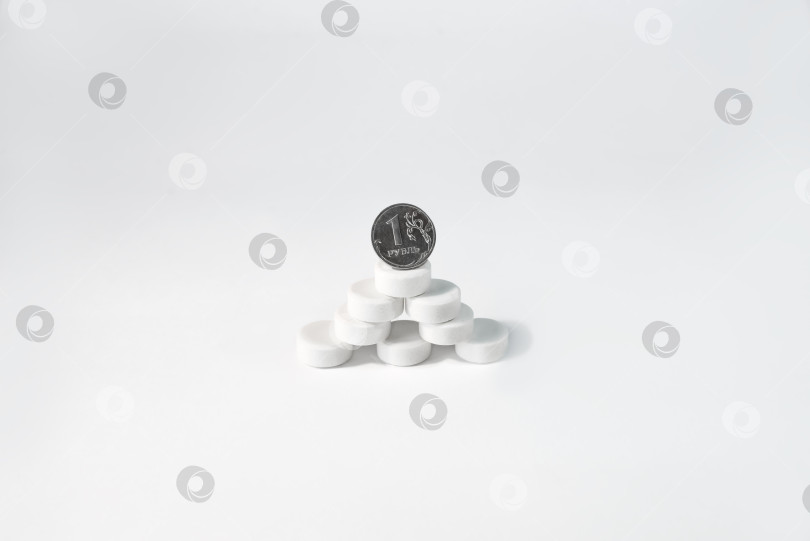 Скачать Монета стоит на горе табличек на белом фоне. Пирамида таблеток и один рубль. Медицина и бюджет здравоохранения, цены на лекарства, расходы на лечение. фотосток Ozero