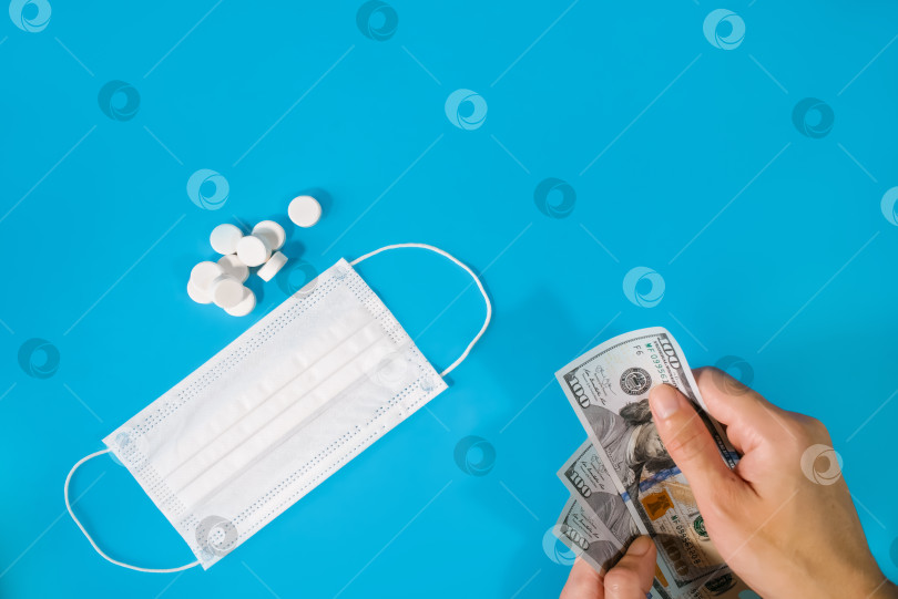 Скачать Мужчина держит в руке деньги на фоне медицинской маски и таблеток. Он символизирует покупку лекарств, стоимость лечения, медицину и здравоохранение, фармацевтический бизнес. фотосток Ozero