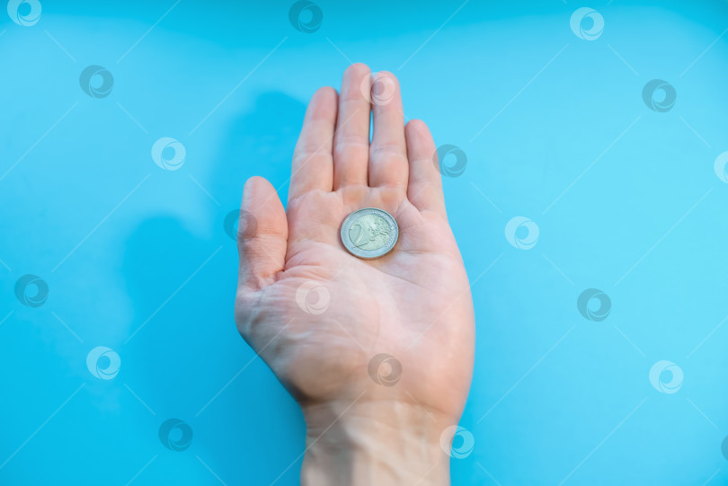 Скачать Мужчина держит на ладони одну монету крупным планом. Два евро в руке, от первого лица. Банкротство, бедность, падение доходов, низкая стоимость. Синий фон фотосток Ozero
