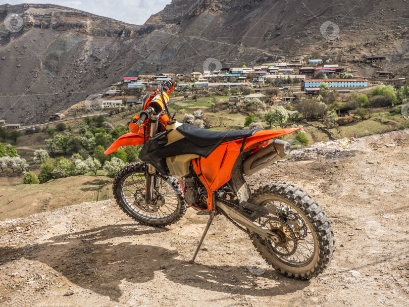 Скачать Оранжевый мотоцикл на утесе на фоне горной деревни и высоких гор. Концепция путешествия. фотосток Ozero